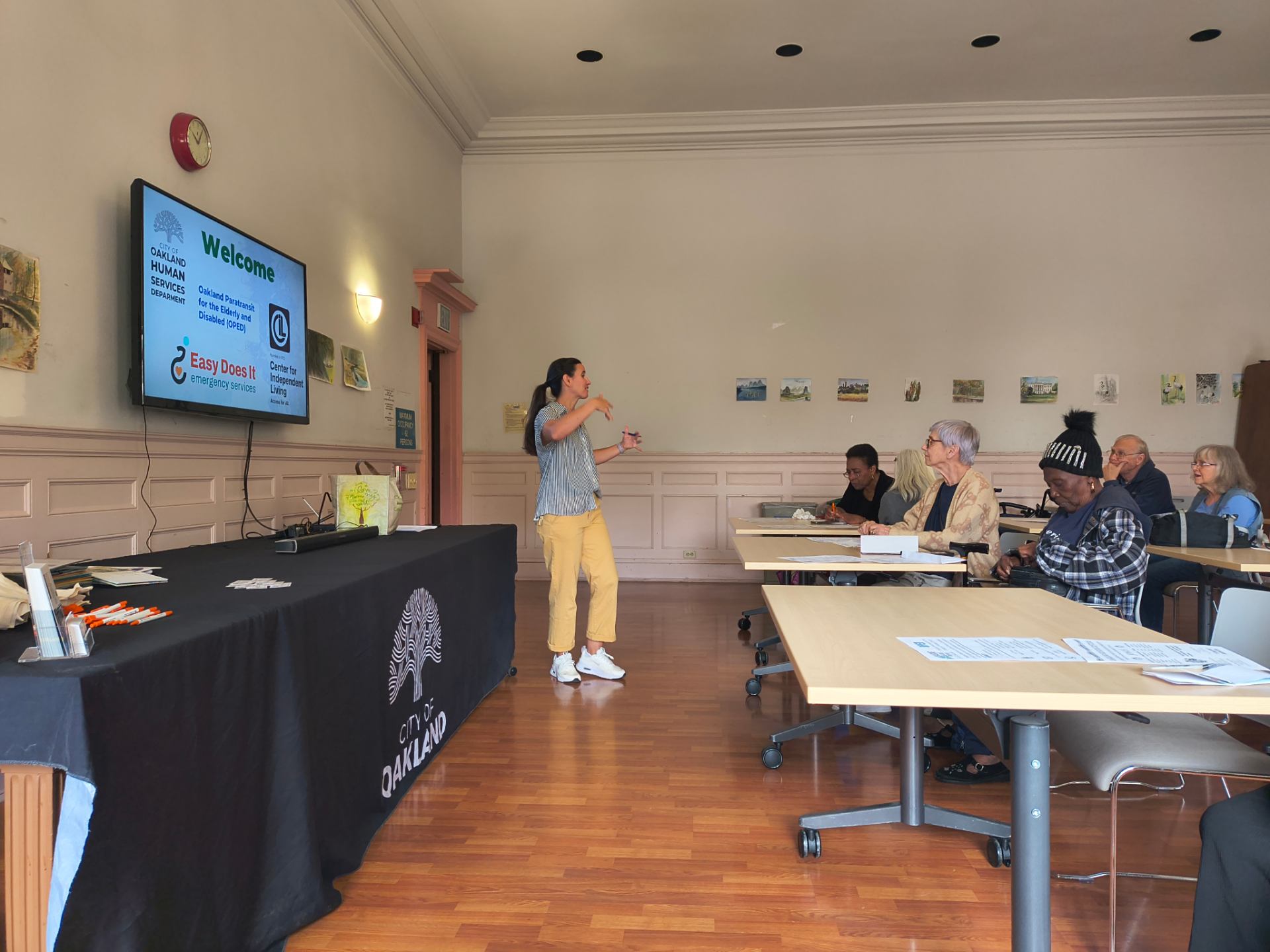 Jessi, Oakland's Aging and Adult Services Coordinator, kicks off our transportation presentation in front of group of seniors. Easy Does It's, The CIL's, and the City of Oakland's logos appear on a TV behind her.